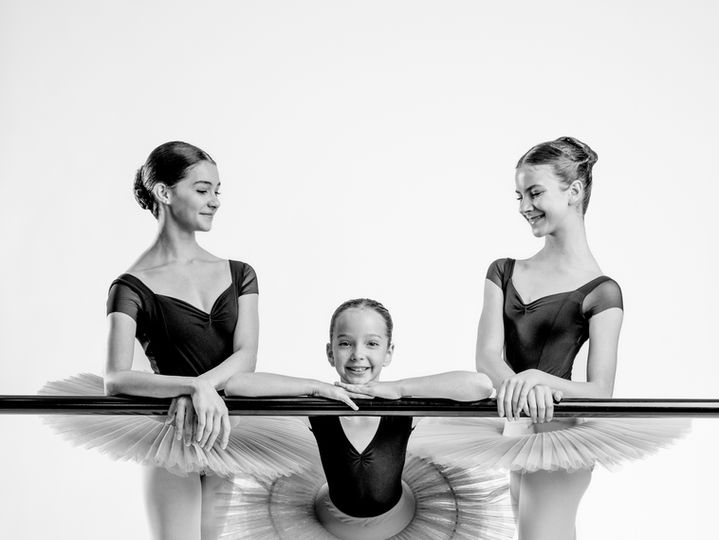 Two ballet students in tutus with a young dancer at the barre